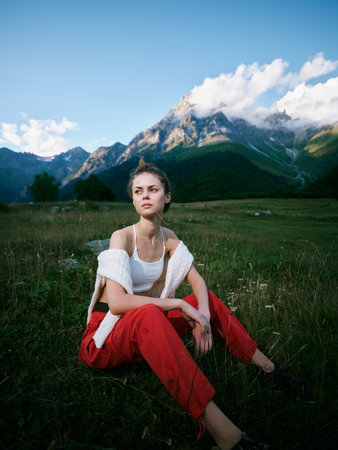 Young woman sits in a grassy meadow with alpine mountains behind, wearing a white top and red pants,...