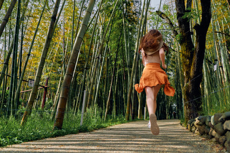 Woman running along a bamboo forest path in an orange dress, motion blur and back view among tall...