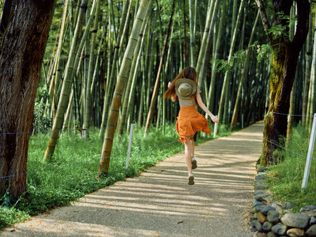 Woman running along a gravel path through a dense bamboo forest wearing a straw hat and orange skirt, enjoying summer nature trail, motion and peaceful green scenery.の写真素材