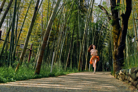 Woman running on a gravel path through a dense bamboo forest, nature trail with sunlight filtering between tall stalks, casual runner with orange jacket tied, motion and freedom.の写真素材