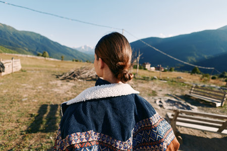 Woman back view in rural countryside wearing patterned jacket, overlooking mountain landscape and valley. Outdoor travel scene with natural light, calm morning and wooden fence.の写真素材