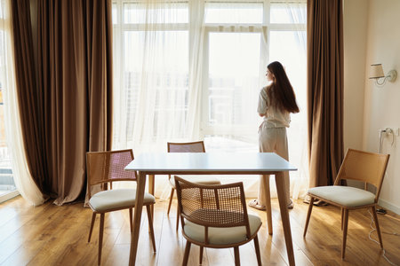Woman by window in morning interior, standing near dining table and chair with curtains, wearing robe and looking outside at sunlight on wooden floor, peaceful home scene of quiet solitude.の写真素材