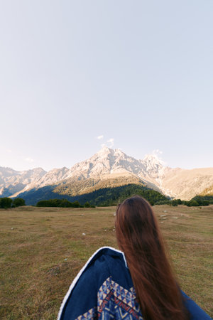 Mountain landscape with woman in meadow, outdoor hiking scene showing long hair and jacket from back, scenic peak and wide sky, travel and adventure mood in natural wilderness.の写真素材