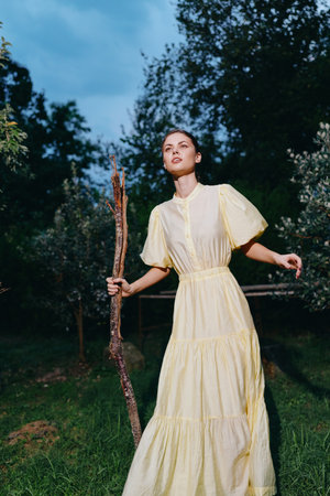 Woman in a pale yellow dress stands in a forest clearing at dusk, holding a wooden staff, exuding calm, ethereal atmosphere and a connection to natureの写真素材