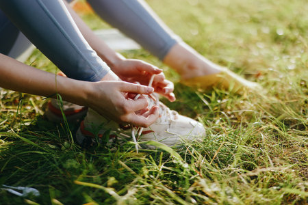 Young woman tying shoelaces on running shoes in a sunny park, representing an active lifestyle with nature background and bright colors, perfect for fitness themesの写真素材
