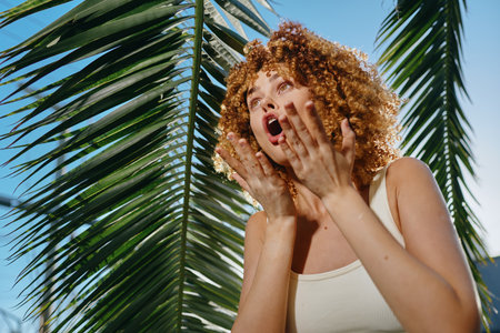 curly woman shouting outdoors with palm leaves background, natural light, casual white tank top, expressive emotion, sunny summer day, vibrant green foliageの写真素材