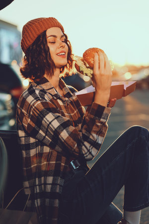 Woman eating a burger outdoors at golden hour, wearing a beanie and plaid shirt, smiling on a street bench with sunlight. Authenticity, candid lifestyle, golden hour glow, mindful living, emotionalの写真素材