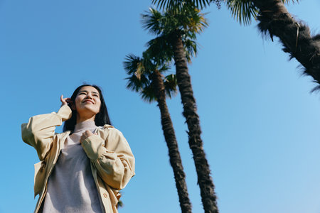 Woman posing under tall palm trees against a bright blue sky, enjoying sunshine in a casual jacket, relaxed mood, outdoor scenery, carefree fashion, confident stance, summer vibeの写真素材