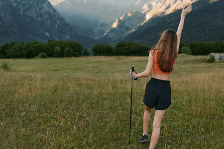 A lone hiker woman traverses a wide meadow framed by distant mountains. She wears a sleeveless top and shorts, raises one hand, and enjoys the fresh outdoor scenery.の写真素材