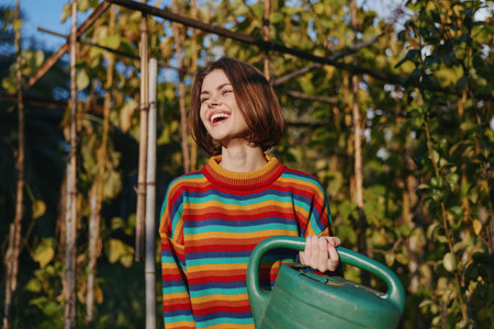 Young woman mid 20s in a striped sweater holding a watering can in a sunny garden by a wooden trellis, smiling and laughing in a casual outfit for lifestyle gardening.の写真素材