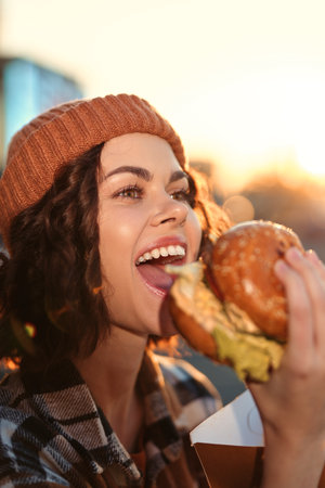 Woman eating burger with big smile in candid beanie outdoor lifestyle portrait during golden hour glow; authenticity and emotional storytelling capture mindful living and urban joy.の写真素材