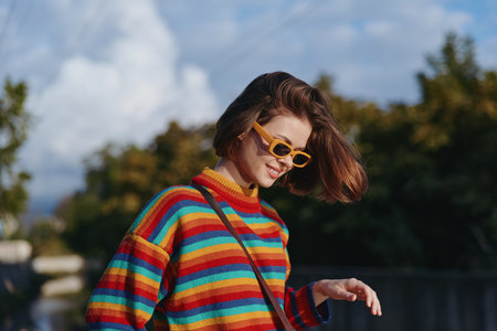 Woman in a vibrant striped sweater and yellow sunglasses walking outdoors, smiling and relaxed, casual outfit with shoulder bag in urban park lifestyle fashion travel sceneの写真素材