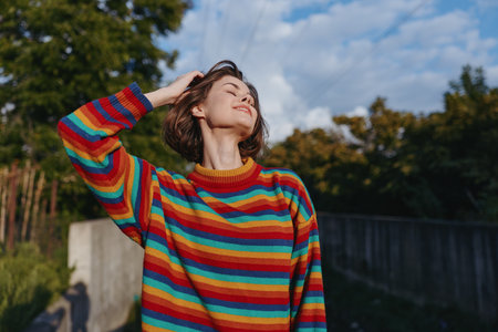 Woman in rainbow sweater outdoors, portrait in sunlight with eyes closed and hand in hair, happy and relaxed young female enjoying nature near concrete wall and trees.の写真素材