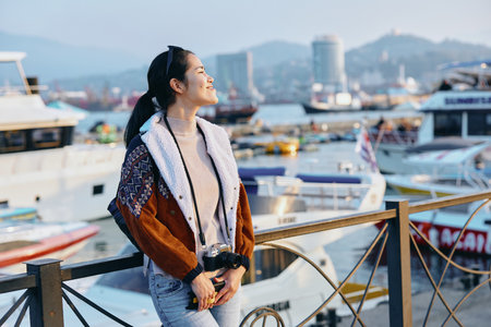 Woman in a warm coat with scarf stands on a wooden pier beside a bustling harbor, boats in the background, clear sky and distant cityscape create a relaxed travel mood by the waterの写真素材