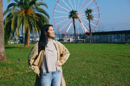 Woman in a beige cape and casual sweater stands in a sunny park with a ferris wheel in the background, enjoying outdoor leisure, relaxed pose, confident style, and fresh air.の写真素材