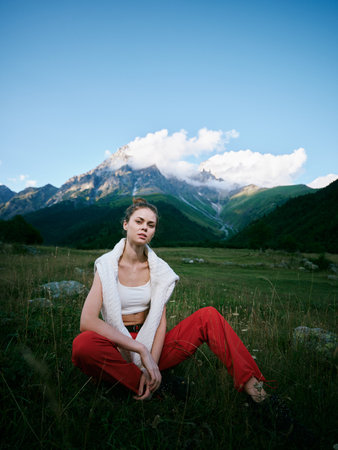Woman in red pants and white top sits in a meadow before towering mountains, highlighting outdoor fashion, nature, and tranquil scenery.の写真素材
