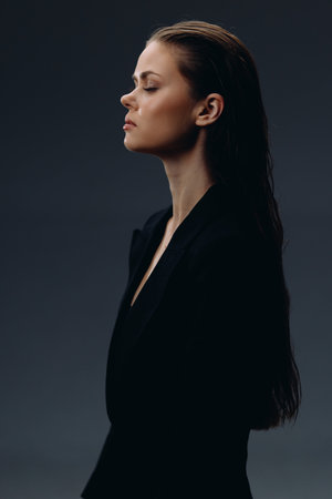 Serious woman with long hair in black outfit standing sideways against dark background, showing calm and determination.の写真素材
