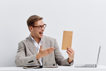 Cheerful young man wearing glasses and stylish gray blazer holding a brown envelope while sitting at a desk with a laptop isolated on white background studio portrait. People lifestyle conceptの写真素材