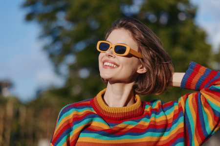Woman smile sunglasses colorful sweater portrait outdoors, happy fashion young female enjoying sunlight and vibrant style, casual knitted look with positive mood and relaxed confident pose.の写真素材