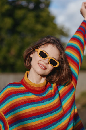 Woman smiling in a colorful striped sweater and yellow sunglasses, short hair, raising her arm outdoors in a sunny park setting. Casual fashion, joyful young lifestyle portrait.の写真素材