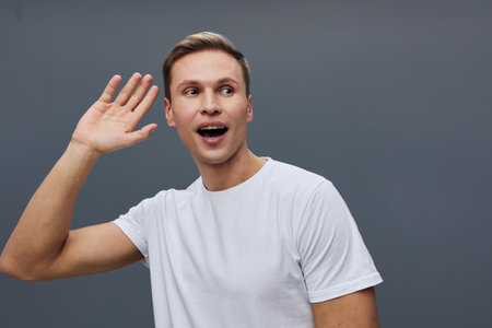 happy young caucasian man wear white t-shirt listen with hand near ear front dark gray background...