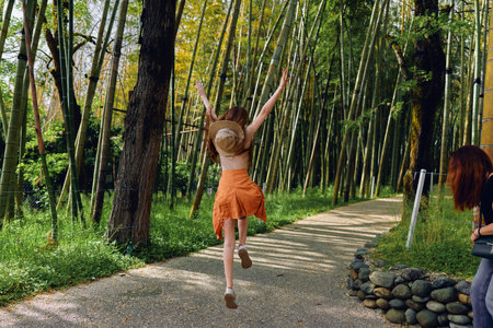 woman jump on a gravel path through a sunlit bamboo forest, orange skirt and straw hat visible as she raises her arms in carefree motion and joyful summer outdoor travel sceneの写真素材
