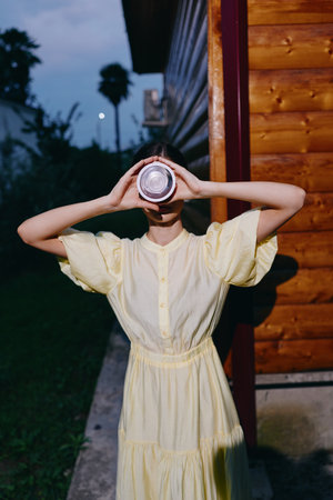 Young woman in a pale yellow dress holds a camera lens to her face outdoors near a wooden cabin at dusk, creating a playful mysterious portrait with moody lightの写真素材