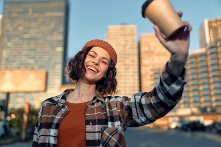 Woman holding coffee cup in urban street, smile and candid lifestyle moment with beanie and plaid shirt, sunlight and happiness combine into golden hour glow, authenticity and mindful living.の写真素材