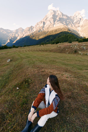 Woman in autumn sweater sits in a mountain meadow, nature landscape with high peaks and soft light,...