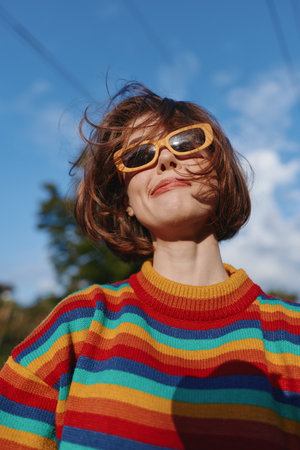 Woman wearing yellow sunglasses and a colorful striped sweater, smiling with short bob haircut outdoors under blue sky. Mid 30s model, playful windblown hair, retro casual outfit for lifestyle andの写真素材