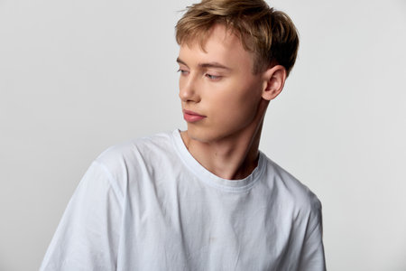 Man in a white shirt gazes pensively to the side in a clean studio setting, minimal background, soft...