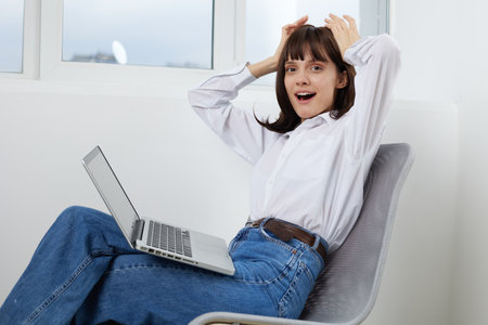 A cheerful young woman sits in a modern home office, laptop on her lap, hands in her hair, and a...