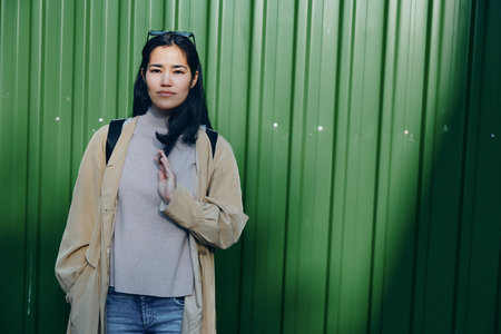 Woman in beige coat and gray sweater stands before a green metal wall, casual outdoor street fashion, confident pose, urban vibe, candid modern style, daytime sceneの写真素材