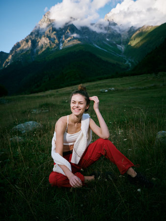 Woman sits on a grassy meadow in a mountain landscape, smiling softly, wearing a white top and red...