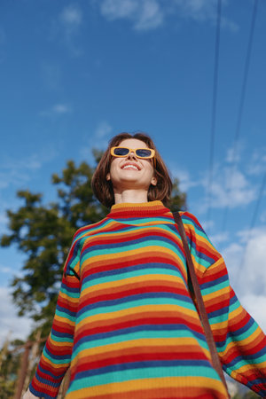 Woman in a colorful striped sweater wearing yellow sunglasses, smiling outdoors under a bright blue sky; young casual outfit with shoulder bag, joyful travel lifestyle portrait.の写真素材