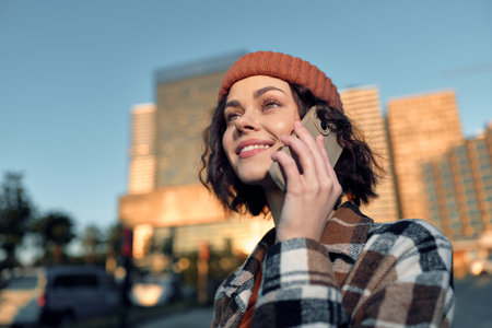 Woman on phone with smile in city, wearing beanie and plaid coat at golden hour glow; candid authenticity and mindful living capture emotional storytelling in modern urban lifestyle.の写真素材