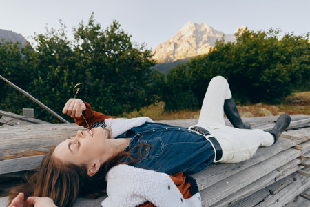 Woman lying on wooden logs eating wild berries in nature by mountain, wearing denim jacket and white pants, relaxed outdoors scene with boots and forest background.の写真素材