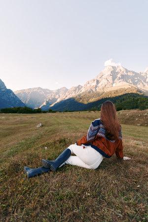 Woman sitting in a meadow gazing at majestic mountains, nature landscape during autumn travel and solitude, cozy sweater and boots on grassy valley with serene outdoor scenery and calm.の写真素材