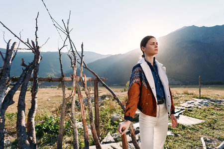Woman in jacket standing in a rural field with mountains behind, sunlight on her face, looking...