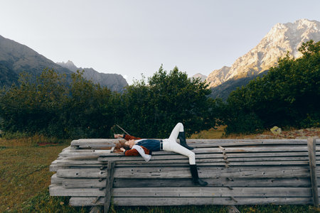 woman lying on wooden bench in mountain nature scene with forest backdrop and meadow, relaxed outdoors lifestyle portrait, casual fashion and peaceful resting in scenic travel escapeの写真素材