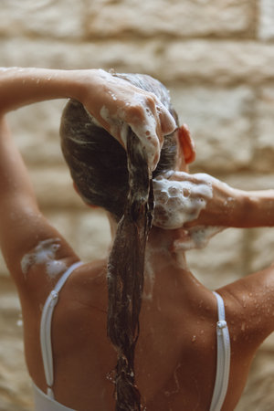 Woman washing long hair with shampoo foam in shower, wet brown hair, bare shoulders, and white straps visible, natural stone wall background creating a warm atmosphere.の写真素材