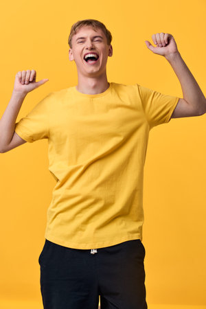 Joyful young man in a yellow shirt celebrates with arms raised, expressing excitement and triumph....