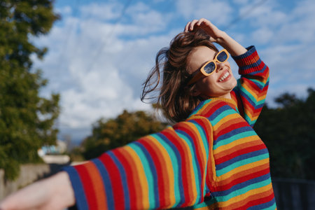 Woman in her mid twenties wearing a rainbow striped sweater and sunglasses, smiling outdoors in an urban park with arms out and wind in hair, joyful casual outfit for travel and lifestyle portrait.の写真素材