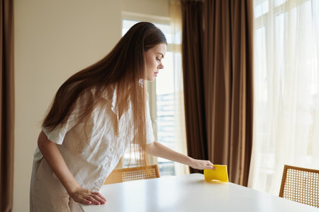 Woman cleaning table with yellow sponge in bright home interior by curtains and sunlight, focused on wiping tabletop for tidy livingroom, domestic routine and care.の写真素材