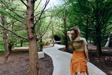 Woman walking on a winding pathway through park trees, back view of a fashionable outfit with orange skirt and heart top, casual summer stroll in green urban forest setting.の写真素材