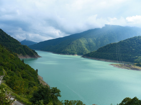 Lake, mountains, forest, reservoir, water, clouds: Turquoise reservoir winding between forested mountains under dramatic cloudy sky. Scenic landscape, remote valley, nature serenity and reflection.の写真素材