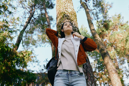 Woman, forest, outdoors, jacket, sweater, backpack, casual, nature, lifestyle, autumn, tall trees frame the scene, sky peeks through leaves, warm sunshine, tranquil mood, exploration and renewal inの写真素材