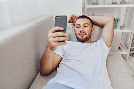 Relaxed man enjoying a moment of leisure while checking his smartphone, wearing casual clothing in a cozy living room setting. Vibrant colors and modern decor enhance the scene.の写真素材