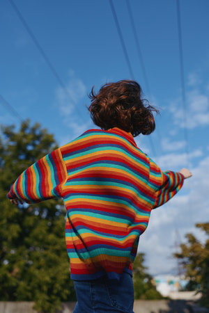Child in rainbow sweater back view with arms outstretched outdoors under blue sky, playful childhood scene in casual clothing and curly hair, motion and joyful freedom.の写真素材