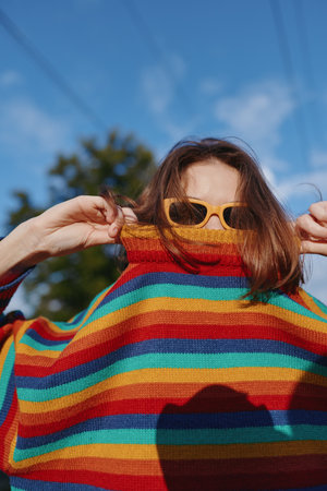 Woman in a rainbow sweater and sunglasses, colorful portrait outdoors on a sunny day. Young woman playful expression, closeup fashion shot with vibrant knitwear and casual style.の写真素材